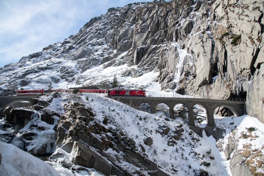 Ein Zug fährt auf einer malerischen Brücke in den Tunnel, umgeben von schneebedeckten Bergen und alpiner Natur.