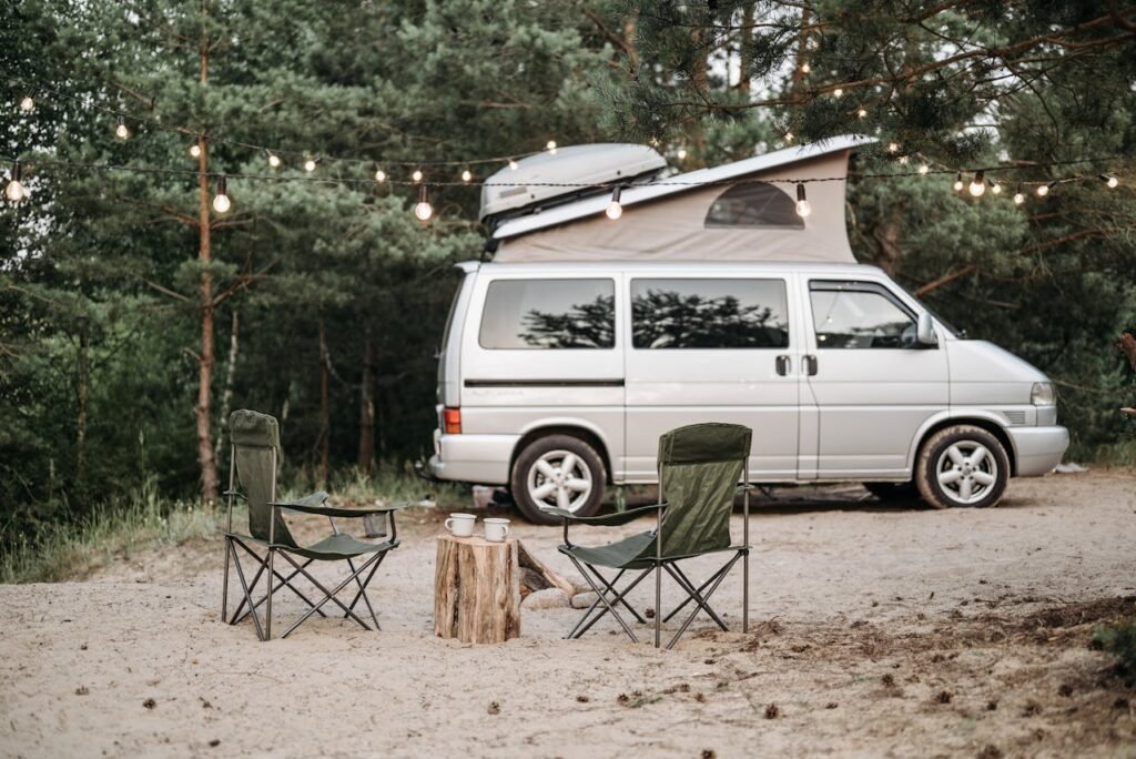 Silberner Festival-Camper mit Aufstelldach auf sandigem Stellplatz. Zwei Campingstühle und Lichterketten schaffen eine gemütliche Ruheoase am Waldrand.