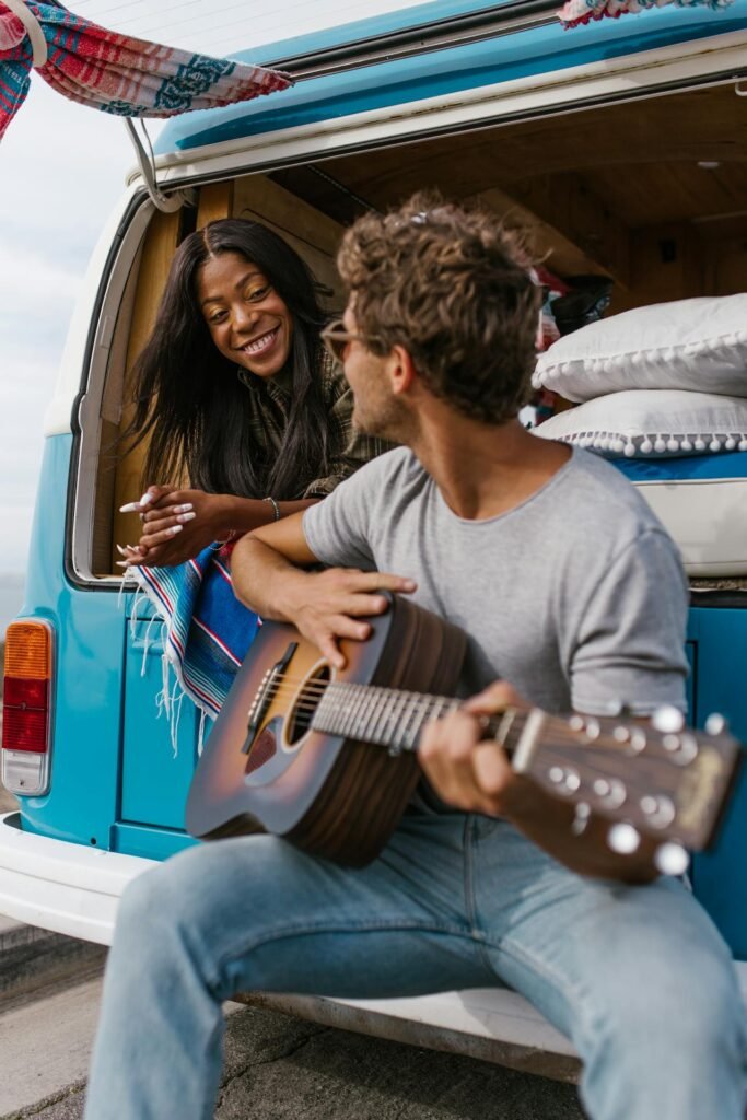 "Junges Paar spielt Gitarre im offenen Heck eines blauen VW-Busses. Fröhliche, entspannte Atmosphäre im Camper-Areal (Festival-Vibe)."