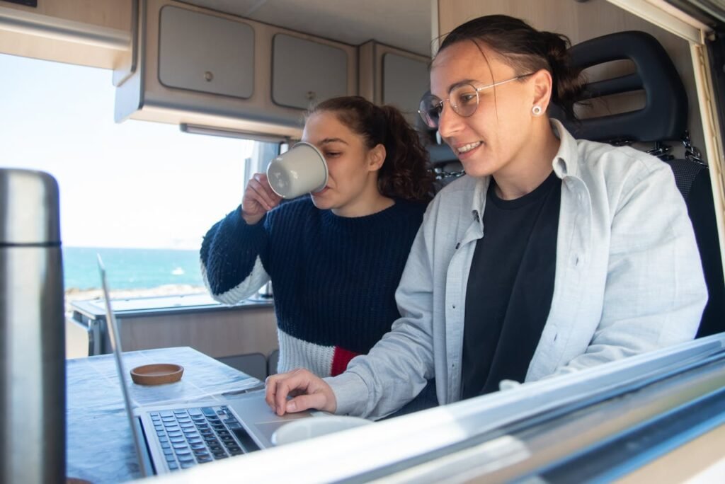 Zwei Frauen arbeiten mit einem Laptop am Tisch in einem Camper Van. Die Szene symbolisiert die Wichtigkeit des Zubehörs und der Gewichtskontrolle bei der Festivalreise. 
Wohnmobil auf Festivals